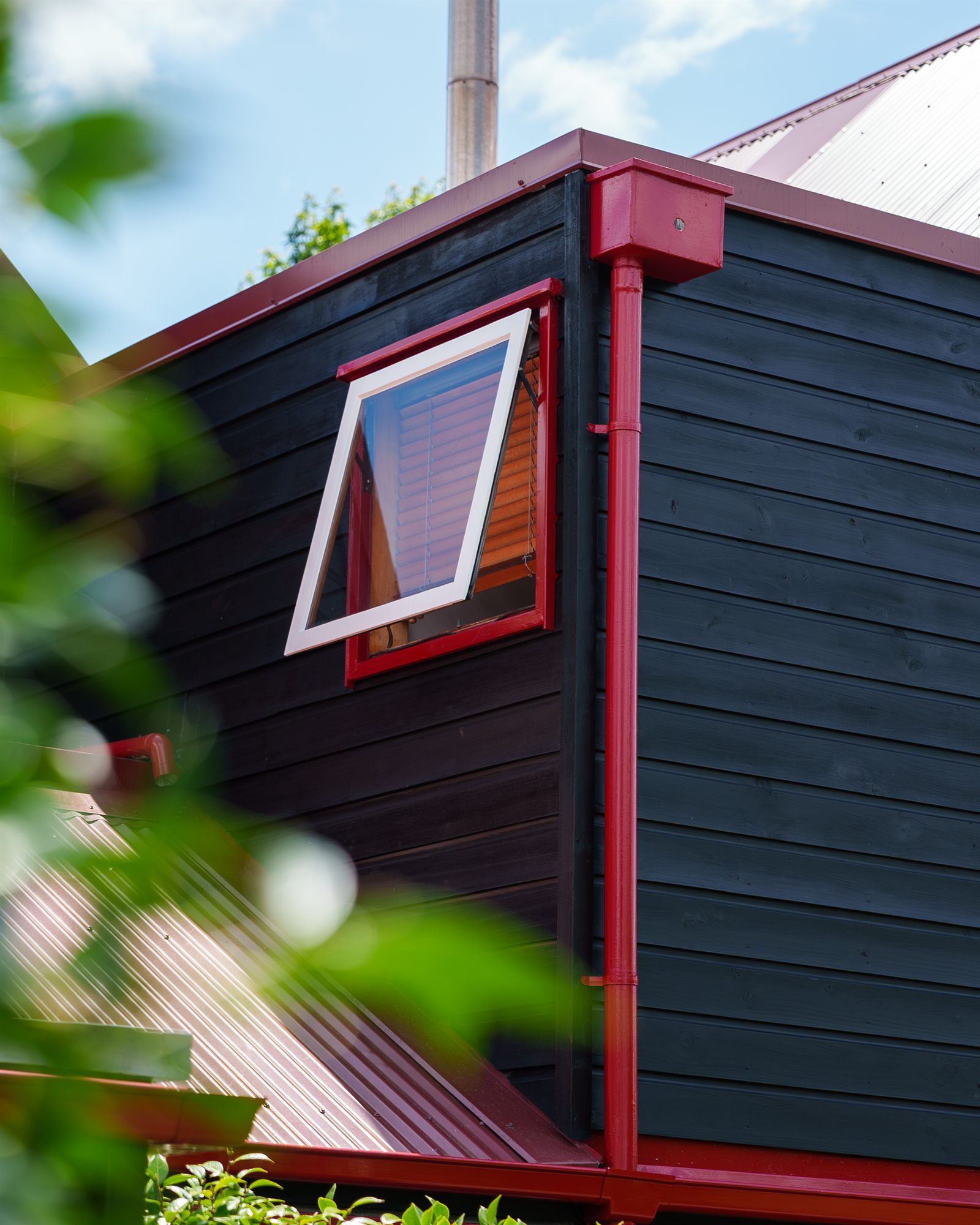 An awning window with a white sash and red reveal, open against black weatherboard, beside a red downpipe and box gutter.