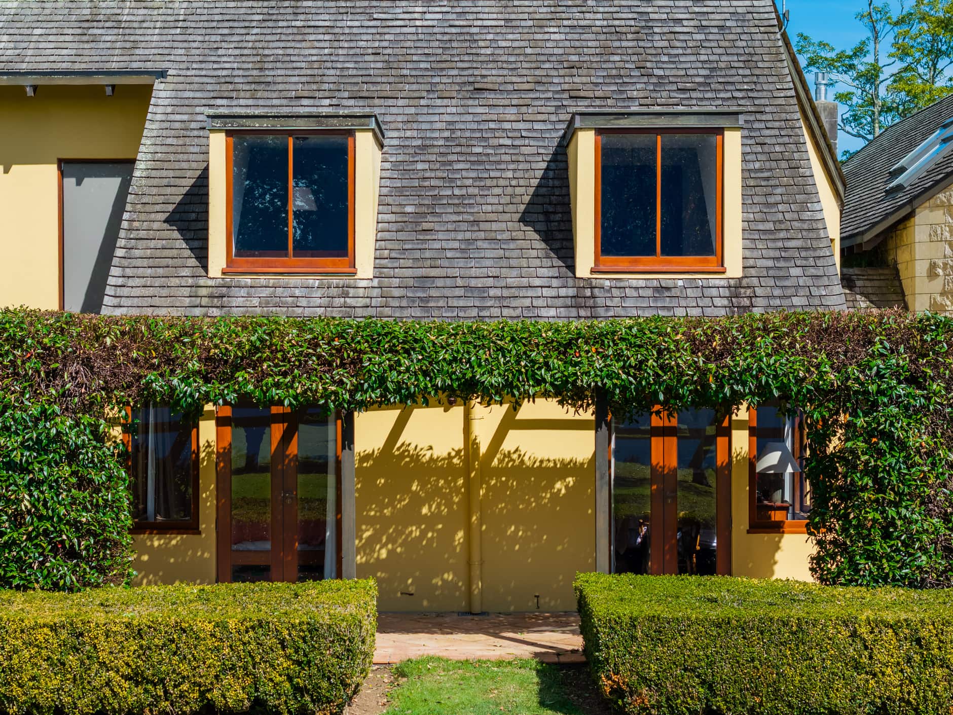 The facade of a cream country house with two dormer windows above a hedge-covered pergola.